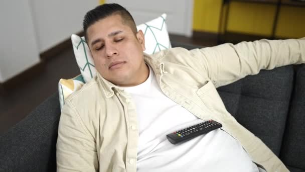 Joven hispano sosteniendo control remoto de televisión acostado en sofá durmiendo en casa - Metraje, vídeo