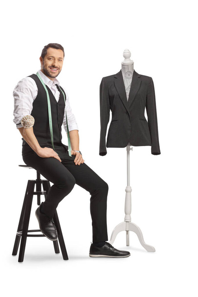 Young tailor with a measuring tape smiling at camera and sitting next to a mannequin doll with a suit isolated on white background - Photo, image