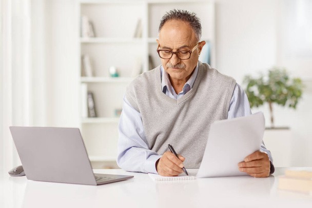Mature man sitting in front of a laptop computer at home and writing a paper document - Foto, Imagen