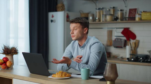 Mosolygó fiatalember elbúcsúzik a webkamerával, amint befejezi a videóhívást a modern konyhában. Vonzó, vidám fickó, aki laptopot húz be, és reggelente croissant kávéval ül az asztalnál. Férfi használ zoom találkozó. - Fotó, kép