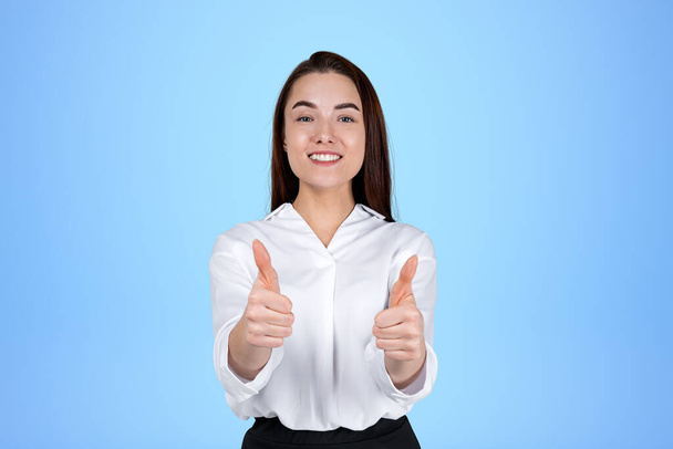 Smiling young businesswoman showing thumbs up, looking at the camera on blue background. Concept of recommend and like. Copy space.  - 写真・画像