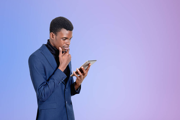 Όμορφος Αφροαμερικανός επιχειρηματίας που φοράει επίσημο κοστούμι στέκεται βλέποντας στο tablet συσκευή κοντά στο άδειο μωβ τοίχο στο παρασκήνιο. Αντίληψη των σύγχρονων gadgets, κινητή επικοινωνία, διαχείριση του χρόνου - Φωτογραφία, εικόνα