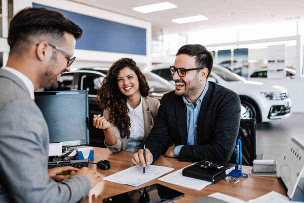 Middle age couple choosing and buying car at car showroom. Car salesman helps them to make right decision. Man signs buyers contract. - Foto, immagini