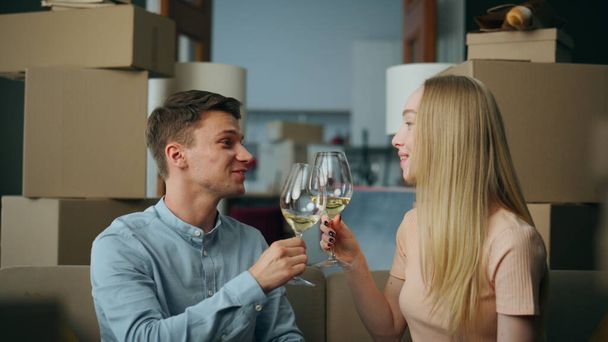 Happy millennials couple celebrating moving into new house drinking wine surrounded packed cardboard boxes close up. Smiling young family clink glasses for beginning new life in own purchased property - Photo, Image
