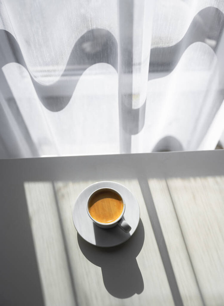 Kubek kawy espresso na stole przy oknie z porannym światłem. Fotografia żywności i napojów - Zdjęcie, obraz