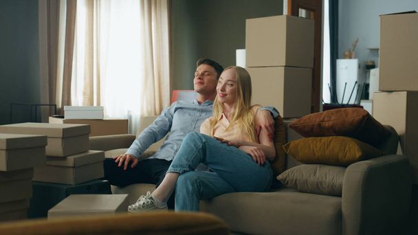 Glückliche Familie, die es sich auf der neuen heimischen Couch gemütlich macht und auf das moderne Wohnzimmer mit gepackten Pappkartons blickt. Zufriedenes junges Paar genießt Einzug in Mietwohnung. - Foto, Bild