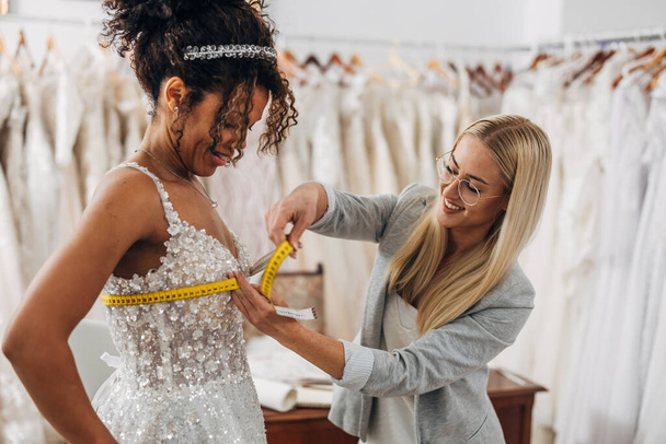A brie in a wedding dress stands in a bridal salon while the tailor takes her measures. - Foto, immagini