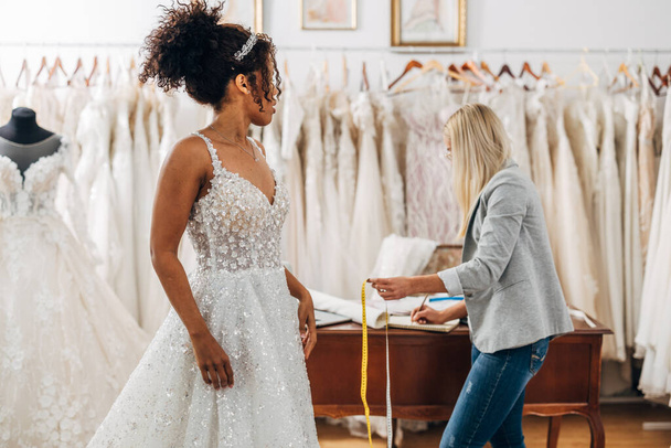 A Multiracial brie in a wedding dress stands in a bridal salon. - Фото, изображение