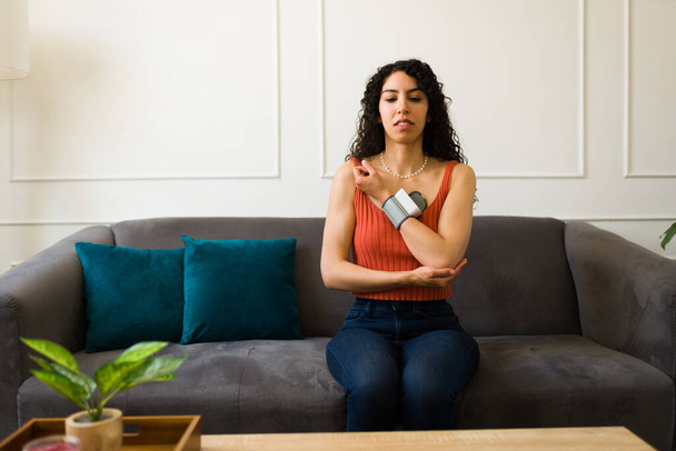 Stressed young woman measuring her blood pressure and having health problems looking sick sitting in the living room - Photo, Image
