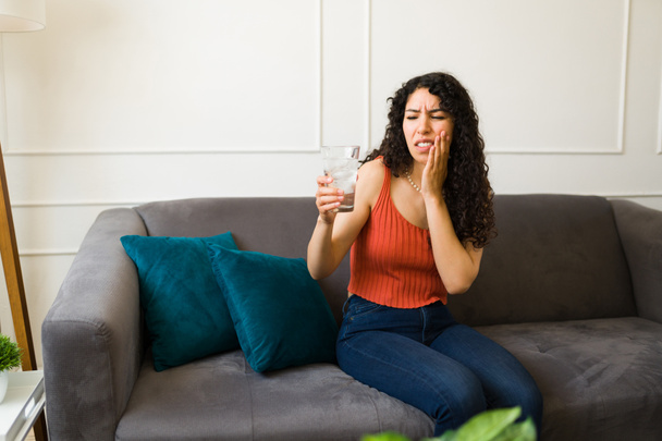 Αναστατωμένη νεαρή γυναίκα που πίνει παγωμένο νερό και υποφέρει από ευαισθησία στα δόντια ενώ κάθεται στον καναπέ στο σαλόνι - Φωτογραφία, εικόνα