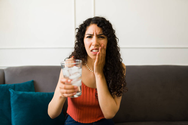 Mujer latina enferma con dolor que sufre de sensibilidad en los dientes y que toca el lado de su cara después de beber agua helada - Foto, imagen