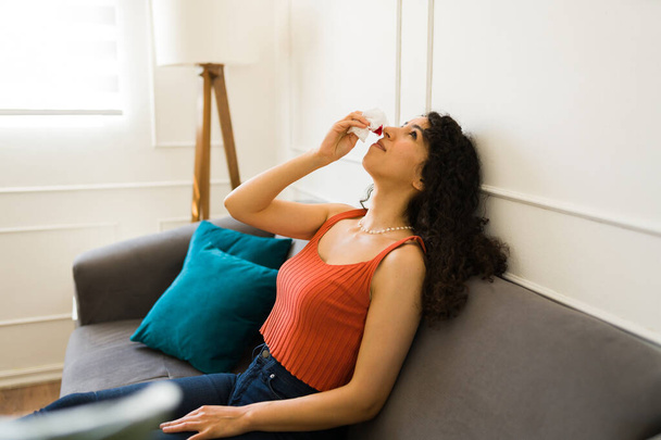 Giovane donna stanca che si riposa sul divano rimettendo la testa indietro con un naso sanguinante e usando un tessuto per il suo sangue dal naso - Foto, immagini
