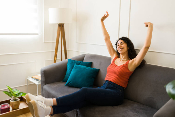Расслабленная красивая латинская женщина поднимает руки и расслабляется, отдыхая на диване в своей новой гостиной - Фото, изображение