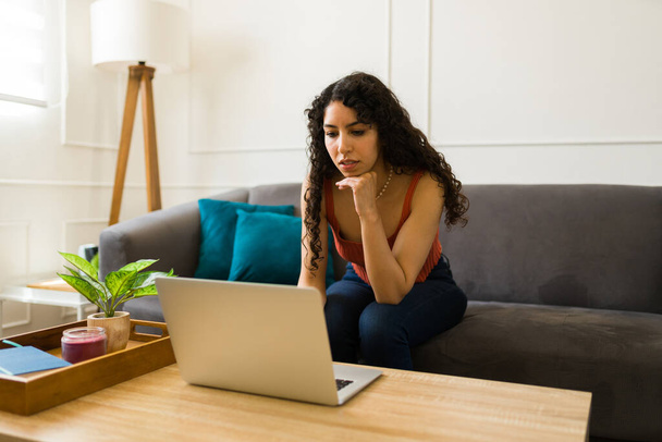 Kiireinen latino nuori nainen kirjoittamalla kannettavan tietokoneen ja työskentelevät kotoa tai tehdä verkko-ostoksia istuu olohuoneessa  - Valokuva, kuva