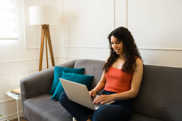 Веселая красивая молодая женщина печатает на ноутбуке, расслабляясь на диване и выходя в интернет - Фото, изображение
