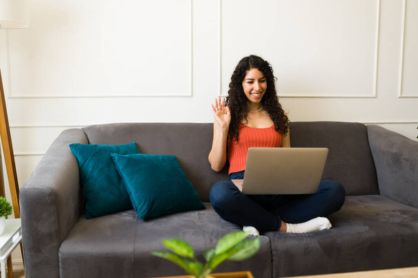 Mujer joven y relajada en el sofá en casa haciendo una videollamada usando su computadora portátil y saludando a amigos o familiares - Foto, Imagen
