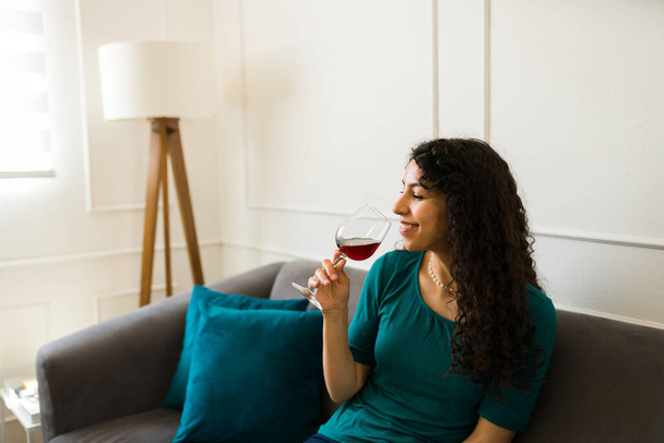 Šťastný hispánský mladá žena relaxační a těší pití sklenice vína, zatímco sám usměvavý odpočívá v obývacím pokoji - Fotografie, Obrázek