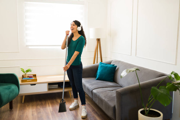 Mujer hermosa hispana haciendo tareas de limpieza barriendo el piso de la sala de estar mientras canta escuchando música con auriculares  - Foto, imagen