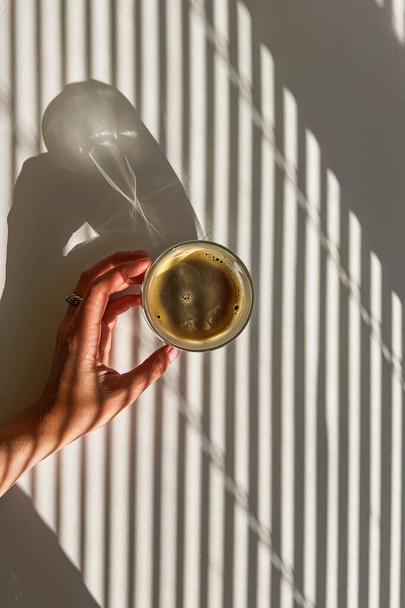 Donna che tiene una tazza di caffè fresco sul tavolo bianco, concetto del mattino, luce naturale e ombre - Foto, immagini