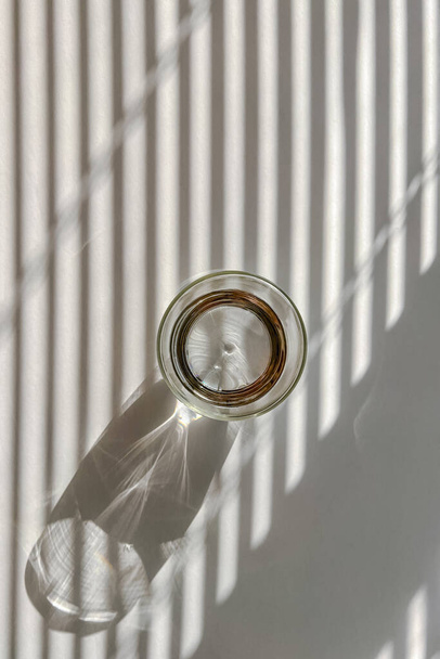 Glass of water on the white table with shadows from the morning light throught the blinds, top view - Фото, зображення