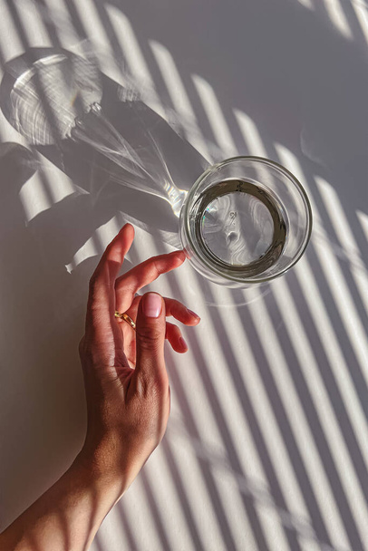 Female hand gently touching a glass of clear water, view from the top, abstract shadows, morning light - Foto, Bild