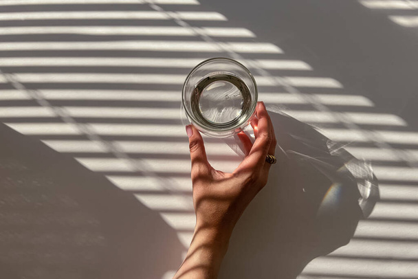 Weibliche Hand hält ein Glas Wasser auf dem weißen Tisch, Blick von oben. Abstrakte Schatten von den Jalousien, Morgenlicht - Foto, Bild