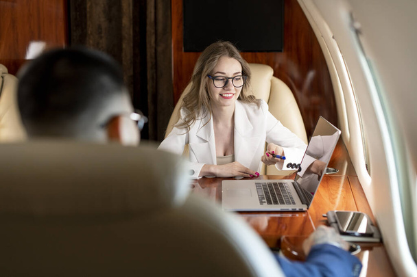 zakelijke bijeenkomst op vliegtuig, zakenvrouw in pak en bril bespreekt omgaan met klant en wijst naar laptop scherm, zakenmensen vliegen in prive-jet en werk - Foto, afbeelding