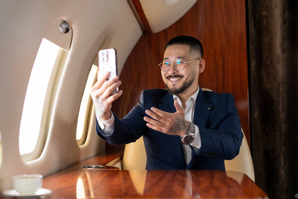 young asian businessman is sitting in private luxury jet and communicate via smartphone video call, korean manager financier in suit and glasses talking on the phone online video broadcast - Photo, Image