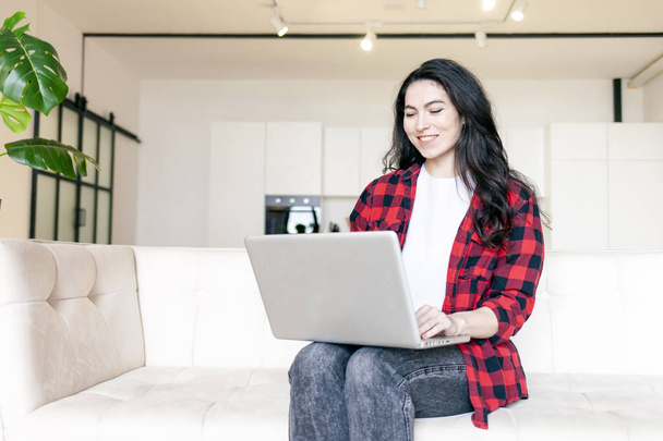 кудрявая женщина в рубашке сидит дома на диване и пользуется ноутбуком, девушка печатает на компьютере и улыбается - Фото, изображение