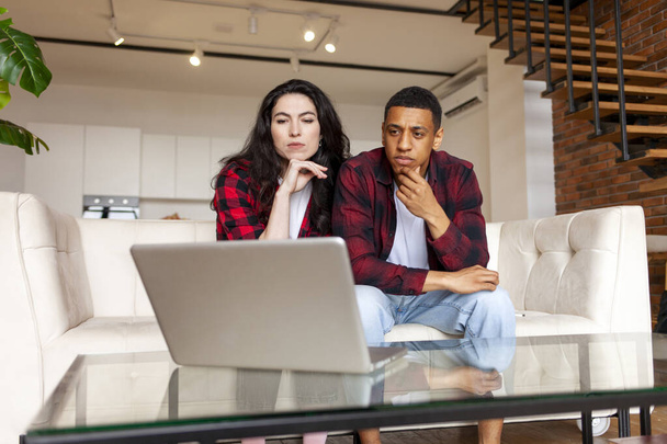 young multiracial couple looking puzzled and serious at laptop on sofa, african american man with european woman confused looking at computer, interracial family using laptop at home - Foto, imagen