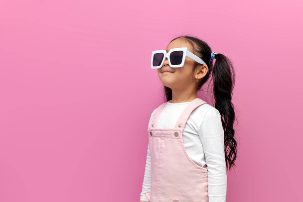 dziewczynka w wieku przedszkolnym w białych okularach przeciwsłonecznych i różowej sukience na różowym odizolowanym tle, dziecko w okularach odwraca wzrok na przestrzeni kopii - Zdjęcie, obraz