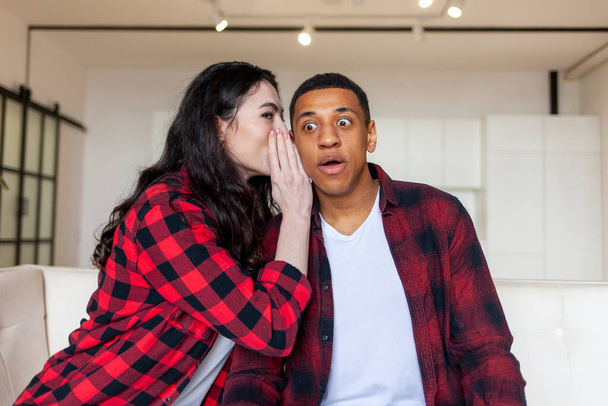 young european woman telling secret to african american man, wife whispering in ear to husband and hinting, shocked and surprised man listening to his wife sitting on sofa at home - Zdjęcie, obraz