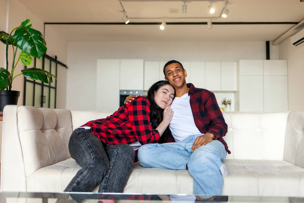 joven feliz pareja multirracial sentado en el sofá en casa y descansando, chico afroamericano con chica sentada en un sofá cómodo, acogedor y concepto de comodidad - Foto, Imagen