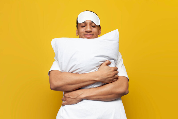 young african american guy in pajamas and sleep mask hugs soft comfortable pillow and smiles on yellow isolated background, the man sleeps and sees good pleasant dreams - Zdjęcie, obraz