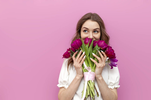 cute young woman in festive outfit holds bouquet of pink tulips and smells them on pink isolated background, girl with flowers for the holiday of March 8 - Photo, Image