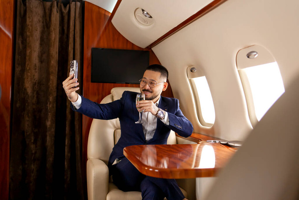 Takım elbiseli Asyalı işadamı ve şampanya bardaklı bardaklar lüks jette uçuyor ve akıllı telefon, Koreli girişimci filmlerini telefonla kullanıyor ve video aracılığıyla iletişim kuruyor. - Fotoğraf, Görsel