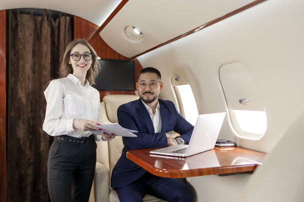 Biznesmeni latają prywatnym samolotem, odnoszący sukcesy azjatycki biznesmen w garniturze z asystentką kobiety z laptopem, a dokumenty latają samolotem i patrzą w kamerę - Zdjęcie, obraz