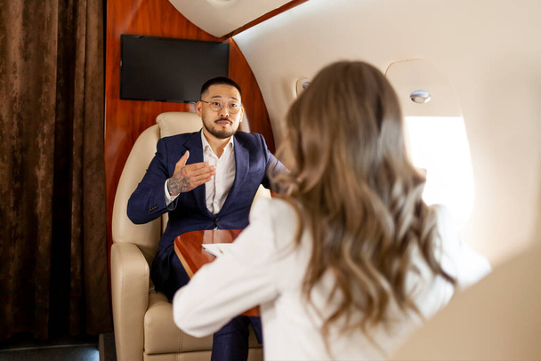 successful asian businessman in suit at meeting with businesswoman partner in private jet, korean manager financier discusses work and plans with colleague in plane - Photo, Image