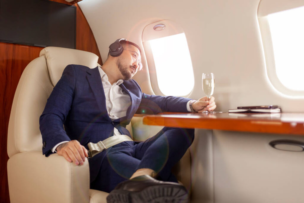 успешный азиатский бизнесмен летает на частном самолете класса люкс с бокалом шампанского и слушает музыку в наушниках, корейский предприниматель в костюме отдыхает и расслабляется в самолете, роскошный образ жизни - Фото, изображение