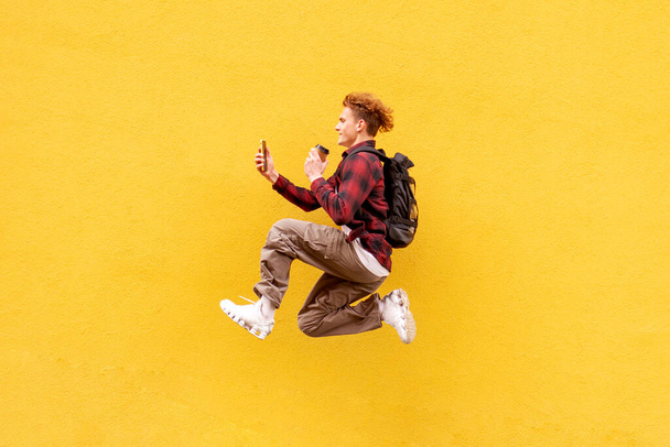 joven estudiante chico alegre con mochila y café utiliza el teléfono y rápidamente se ejecuta en el aire y salta sobre el fondo de la pared aislada amarilla, hombre con teléfono inteligente llega tarde y con prisa - Foto, Imagen