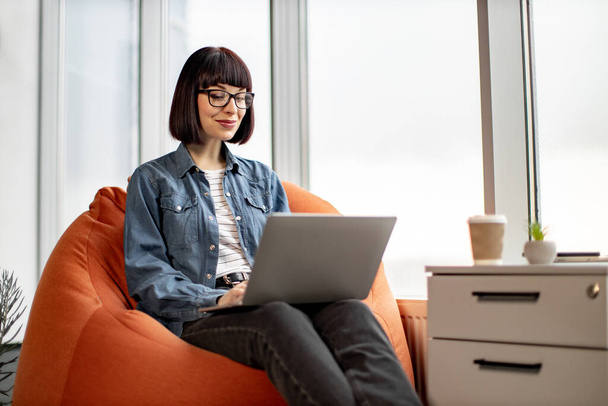 Young pretty female in jeans and jacket having video conference on laptop and spreading hands in glassy office. Attractive short haired woman in spectacles sitting on chair and talking to coworkers. - Fotografie, Obrázek