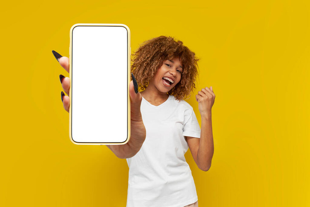 gelukkig krullend amerikaans meisje winnaar toont blanco smartphone scherm en viert overwinning op geel geïsoleerde achtergrond, jonge vrouw adverteert kopiëren ruimte op de telefoon weer te geven en verheugen zich in geluk - Foto, afbeelding