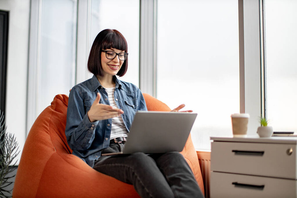 Young pretty female in jeans and jacket having video conference on laptop and spreading hands in glassy office. Attractive short haired woman in spectacles sitting on chair and talking to coworkers. - Fotó, kép