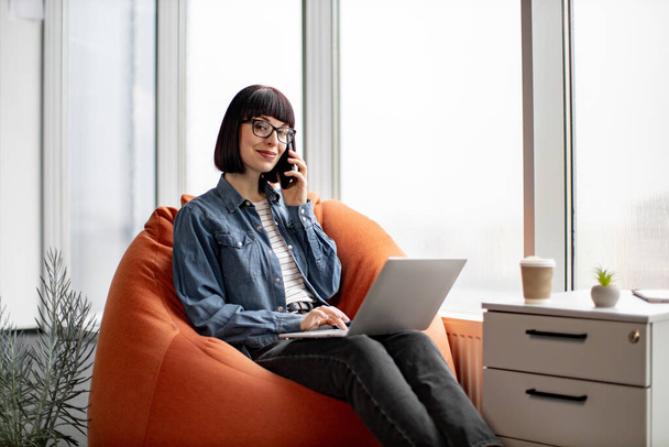Young caucasian lady in casual clothing and glasses speaking on smartphone while working on laptop in office. Smiling woman sitting on orange soft chair and discussing key moments of new project. - Fotoğraf, Görsel