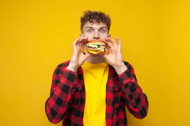 young curly guy student eats a burger on a yellow background, a man with fast food - Foto, afbeelding