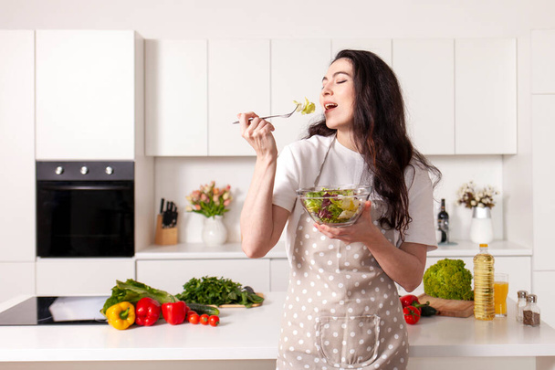 молодая брюнетка в фартуке держит вегетарианский салат на белой современной кухне, домашний повар девушка ест свежий приготовленный салат из тарелки, концепция здоровой пищи - Фото, изображение
