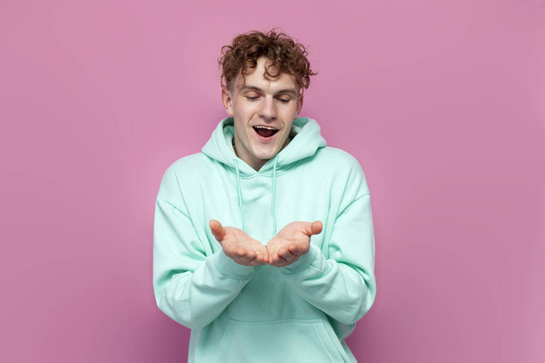 young curly guy in mint hoodie holds empty hands in front of him on pink isolated background, man holds his palms in front of him and looks at his hands - Фото, изображение