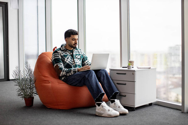 Casually dressed indian programmer working closely on writing and testing codes for new mobile applications in spacious glassy office. Focused man using personal laptop for doing particular job. - 写真・画像