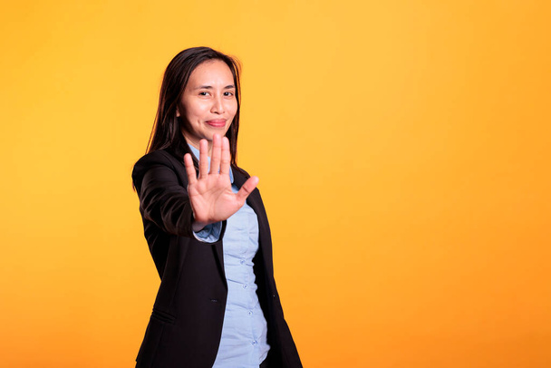 Negativa empresaria filipina haciendo stop gesture con la mano delante de la cámara, posando en estudio sobre fondo amarillo. Mujer seria mostrando gesto de rechazo, expresando rechazo. - Foto, imagen