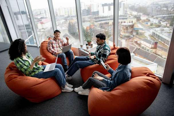 Diverse group of professionals using smart gadgets while resting in circle of soft seats in creative workspace. Multiracial managers in street style clothes planning e-commerce investment strategy. - Valokuva, kuva